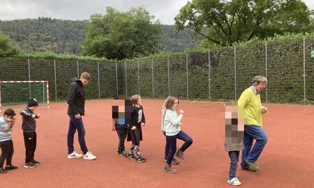 Die Musiklehrerin läuft ganz vorne, ihr folgen sieben Kinder und der Musiklehrer. Alle laufen mit den Füßen rhythmisch auftretend in einer Reihe hintereinander über den Sportplatz.