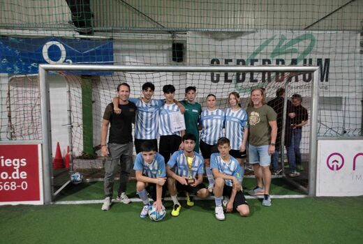 Acht Schüler in Fußballtrikots befinden sich in zwei Reihen vor einem Tor. Die hintere Reihe steht, vorne knien drei Schüler. Vorne hält ein Junge einen Pokal. Rechts und links hinten steht jeweils ein Lehrer..