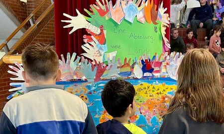 Zwei Jungen und ein Mädchen stehen vor einer Weltkarte mit einem Rahmen aus bunt gestalteten Händen und der Überschrift: Eine Abteilung - viele Länder. Im Hintergrund sitzen viele weitere Kinder auf den Stufen in der Aula.