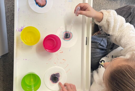 Aus der Vogelperspektive sieht man drei Kinder, die vor einem weißem Tablett sitzen, zwei Kinder tropfen mit einer Pipette Wasser auf ein zum Teil bunt gefärbtes Papier, ein Kind zeigt auf sein Papier, auf dem Tablett stehen außerdem drei Wasserbecher