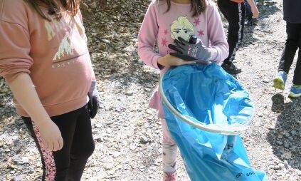 Zwei Drittklässlerinnen stehen mit Handschuhen und Mülltüte vorne, während im Hintergrund weitere Drittklässler und eine Lehrerin auf der Suche nach Müll über den Waldweg laufen.