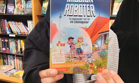 Ein Schüler steht in der Buchhandlung vor einem Bücherregal und zeigt das Buchcover des vorgestellten Buches "Mission Roboter".