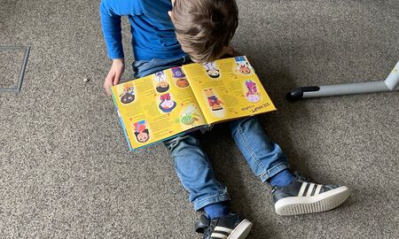 Ein Schüler sitzt auf dem Teppichboden und schaut in ein Buch auf seinem Schoß. Auf der aufgeschlagenen Seite sieht man Menschen mit unterschiedlichem Aussehen.