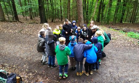 Der Waldpädagoge und die Schülerinnen und Schüler stehen auf einem Waldweg eng beieinander im Kreis und gehen in die Hocke.