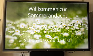 Auf einer digitalen Tafel steht der Schriftzug "Willkommen zur Sommerandacht. Schön, dass du da bist!" Im Hintergrund des Schriftzuges ist eine Wiese mit Gänseblümchen zu sehen.