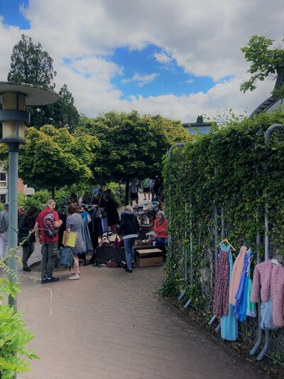 Ein Flohmarktstand mit Kunden, an der Seite hängen zu verkaufende Kleider am Zaun