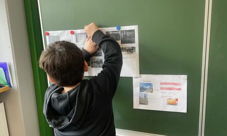 An der Tafel hängt ein DIN A4-Übersichtsblatt über Österreich. Ein Schüler hängt links daneben mit Magneten noch drei Blätter mit Informationen über Serbien auf.