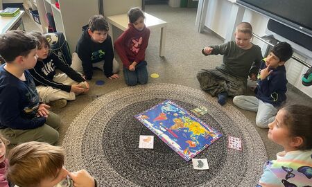 Die Kinder einer ersten Klasse sitzen auf dem Boden in einem Sitzkreis. In der Mitte auf einem runden Teppich liegen eine Weltkarte und kleine Zuordnungskarten.