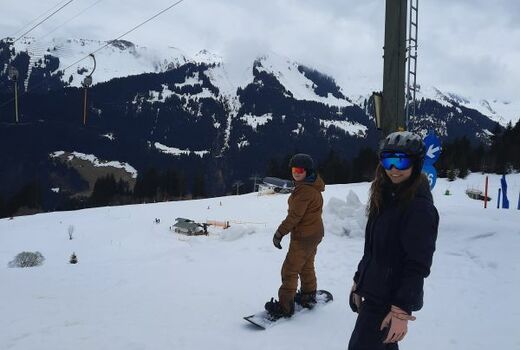 Schneelandschaft mit Bergen im Hintergrund, zwei Jugendliche auf Snowboards stehen bei einem Schlepplift