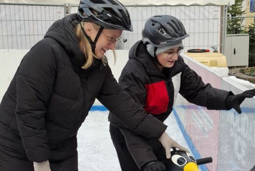 Ein Schüler hebt sich an der Balustrade der Eisbahn. Die Lehrerin hilft.