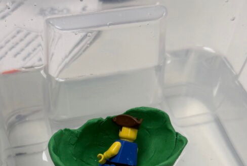 In einem Behälter mit einer Flüssigkeit schwimmt eine kleine grüne Schale, darin liegt eine Lego-Figur