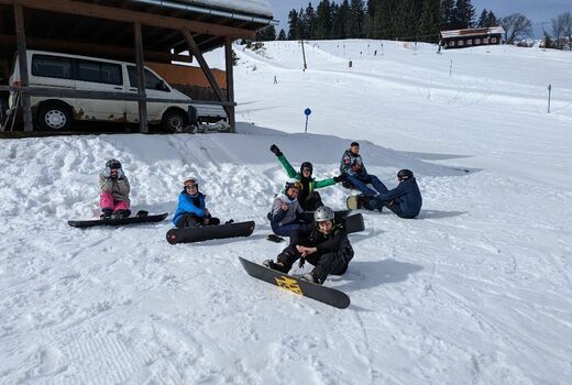 Sieben Jugendliche sitzen mit ihren Snowboards im Schnee. Sie lachen in die Kamera.