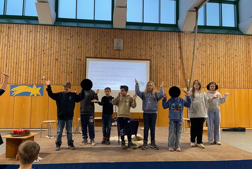 Acht Kinder der vierten Klasse stehen auf der Bühne und beten das Vater Unser vor, sie heben grade ihre Hände nach oben, im Hintergrund ist über den Beamer der Text zu sehen.