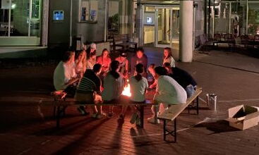 Eine Gruppe Jugendlicher sitzt am Lagerfeuer vor dem Internatsgebäude