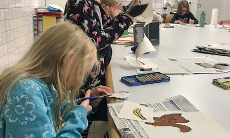 Zwei Schülerinnen schneiden etwas aus Papier aus. Vor Ihnen liegen gemalte Bilder auf Zeitungspapier. Im Hintergrund malen Kinder.