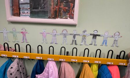 Über einer Garderobe voller Jacken hängen von den Kindern gestaltete Figuren, die sie selbst darstellen sollen. Die Figuren halten sich an den Händen und bilden eine lange Menschenkette.