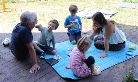 Drei Kinder und zwei Erwachsene sitzen gemeinsam auf einer Decke im Garten und spielen ein Kartenspiel