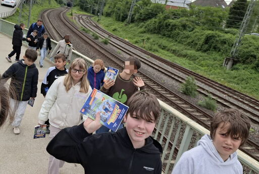Die Kinder sind auf dem Rückweg zur Schule auf einer Brücke über Bahngleisen und halten ihr Buch in die Kamera