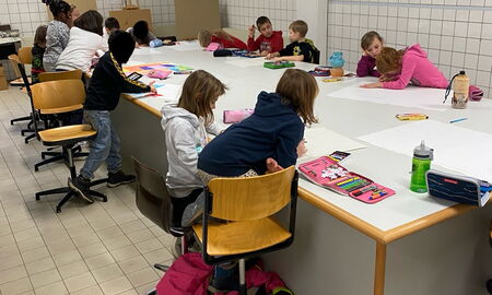 Schüler und Schülerinnen sitzen um einen großen Tisch im Kunstraum und malen zu Zweit an großen Bildern mit Buntstiften.