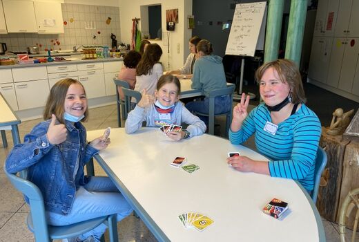 Im Vordergrund sitzen drei lachende Mädchen an einem Tisch und spielen UNO. Im Hintergrund sitzen fünf Mädchen an einem Tisch und spielen.