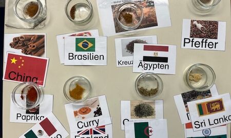 Auf einem Tisch stehen verschiedene Gewürze aus unterschiedlichen Ländern. Die Gewürze sind in Schüsseln, darunter liegen Schilder mit der Bezeichnung und einem Bild sowie den jeweiligen Herkunftsländern mit Flaggen.