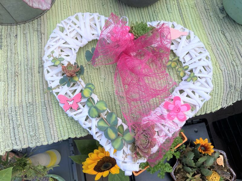 Ein dekoratives Herz aus weißem Korbgeflecht beklebt mit Accessoires in pink und grün