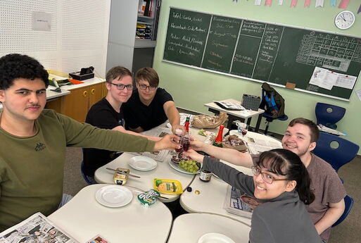5 Schüler*innen sitzen an einem Frühstückstisch und stoßen an