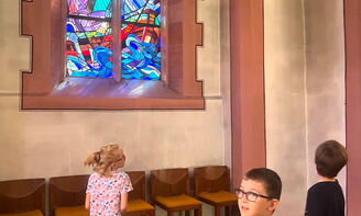 Drei Kinder entdecken die Kirche. In der Mitte des Fotos leuchtet ein buntes Kirchenfenster, auf dem die Geschichte der Sturmstillung abgebildet ist