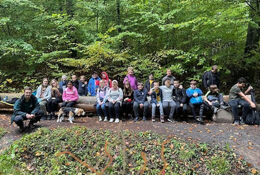 21 SchülerInnen, eine Lehrerin und der Förster mit seinem Hund posieren für ein Gruppenfoto im Wald. Die SchülerInnen sitzen auf einem langen Baumstamm.