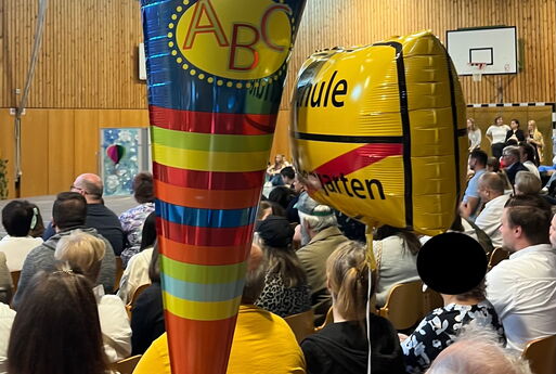 Die Gäste der Einschulungsfeier sitzen in der Sporthalle und in der Mitte sind zwei große Luftballons in Form einer Schultüte und eines Städteschildes auf dem Schule steht und Kindergarten durchgestrichen.