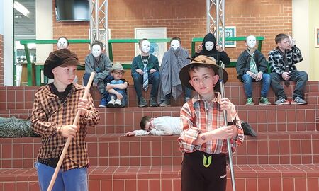 Zwei Kinder stehen als Hirten verkleidet vorne. Im Hintergrund sitzen sieben Kinder mit weißen Masken und weitere Kinder liegen auf dem Boden.