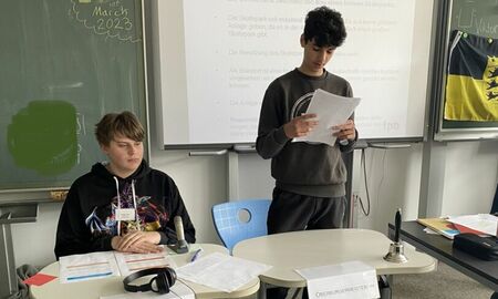 Zwei Schüler sind vor der Tafel bzw. dem Whiteboard. Einer sitzt und einer steht. Der stehende Schüler liest etwas von einem Blatt vor. Hinter ihm am Board steht "Beschlussvorlage".