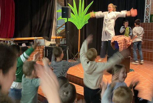 Eine Lehrerin springt rechts im Bild mit beiden Armen ausgestreckt in die Luft auf einer Bühne, links im Bild stehen viele Kinder vor ihr und springen ebenfalls in die Luft