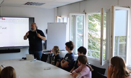 Kinder schauen bei einem Vortrag auf ein Panel. Der Referent steht vorne und erklärt