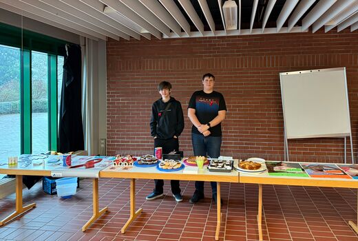 zwei Schüler am Stand in der Aula mit Kuchen und Informationsmaterial