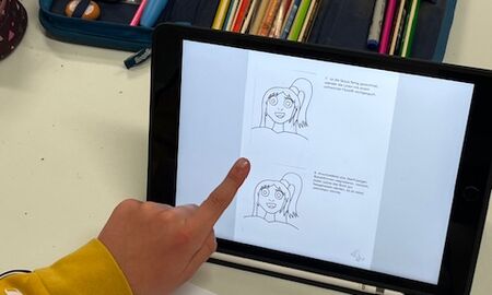 Ein iPad mit einer visuellen Anleitung zum Zeichnen eines Gesichtes steht auf dem Tisch. Davor liegt ein weißes Blatt, auf dem mithilfe der Anleitung ein Gesicht gezeichnet wurde. Ein Kind zeigt gerade mit dem Finger auf das iPad.