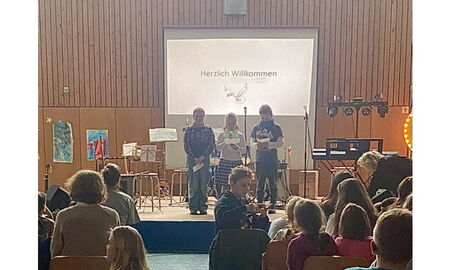 Drei Schülerinnen und Schüler der Klasse drei stehen auf der Bühne mit Texten und Mikrofonen in der Hand. Im Hintergrund auf der Leinwand steht "Herzlich Willkommen". Darunter ist ein Bild von einer Friedenstaube.