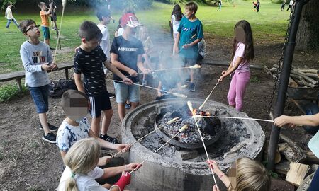 Schülerinnen und Schüler stehen und sitzen mit Stockbrotstöcken um eine Feuerstelle herum, die restlichen Kinder spielen auf der Wiese im Hintergrund