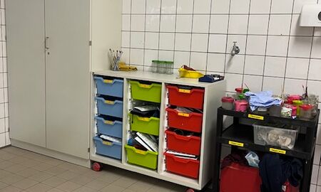 In einer Ecke des gefliesten Kunstraumes stehen links ein großer Schrank, daneben ein Rollcontainer mit bunten Schubladen und rechts ein Tisch zur Aufbewahrung der Materialien.