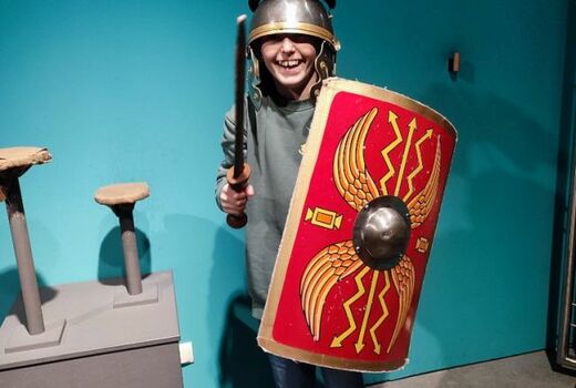 Ein Schüler ist als römischer Soldat verkleidet. Der Helm verdeckt das halbe Gesicht. Er lach und hält sein Gladius in die Höhe.