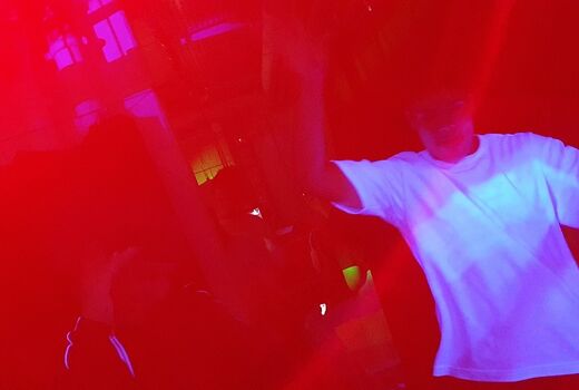 Zu sehen sind rote Lichteffekte und ein Junge im weißen T-Shirt. Er tanzt.