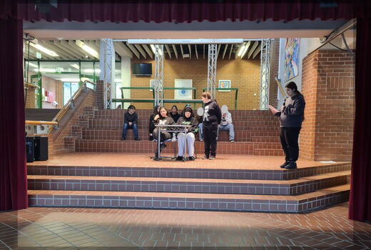 Vier Schüler sind auf der Buehne, zwei sitzen am Tisch, einer steht daneben und seitlich liest ein stehendes Mädchen etwas von einem Papier ab. Sie hat ein Mikrofon in der Hand. Hinter der Gruppe sitzen weitere Schüler auf den Stufen.