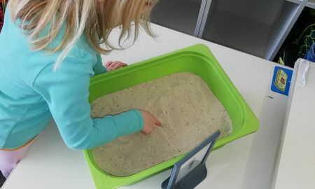 Ein Mädchen schreibt an einer Station den Großbuchstaben M in Sand in einer Kiste.