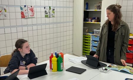 Im Kunstraum steht die Lehrerin am Kopf des Tisches. Links vor ihr sitzt eine Schülerin mit iPad und beide schauen sich an. Auf dem Tisch stehen iPads, Farben und weitere Materialien.