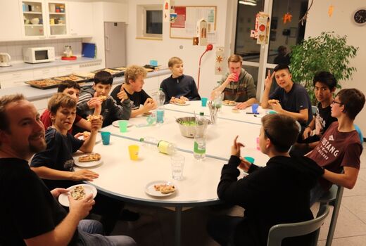 Zehn Jungen und ein Betreuer essen Pizzabrötchen. Sie sitzen gemeinsam an einem ovalen Tisch.