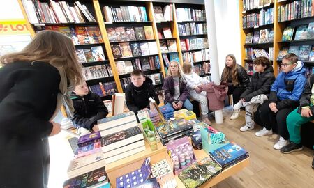 Die Schülerinnen und Schüler sitzen im Halbkreis in der Kinderbuch-Ecke. Die Buchhändlerin zeigt ihnen das Buch "Mission Roboter".