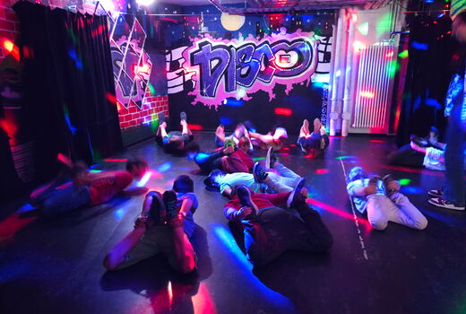 Einige Kinder liegen in einem Raum mit dem Schriftzug "Disco" bei buntem Discolicht auf dem Bauch und halten ihre Füße fest.