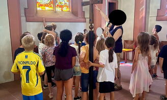 Eine große Kindergruppe steht vor einem bunten Kirchenfenster. Eine Pfarrerin zeigt auf zwei Engel auf dem Fenster.