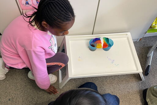 Zwei Kinder sitzen übers Eck an einem weißen Tablett auf dem Boden, auf dem Tablett stehen zwei Becher mit bunt gefärbten Papier darauf