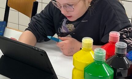 Eine Schülerin sitzt vor einem weißen Blatt und mehreren Flaschen mit Acrylfarbe an einem Tisch. Sie hält einen Füller in der Hand und recherchiert an einem iPad.